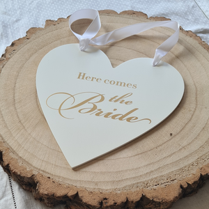 Ivory wooden heart shaped sign with white satin ribbon and gold text that reads, Here comes the Bride. A perfect accessory for pageboy or flowergirl.